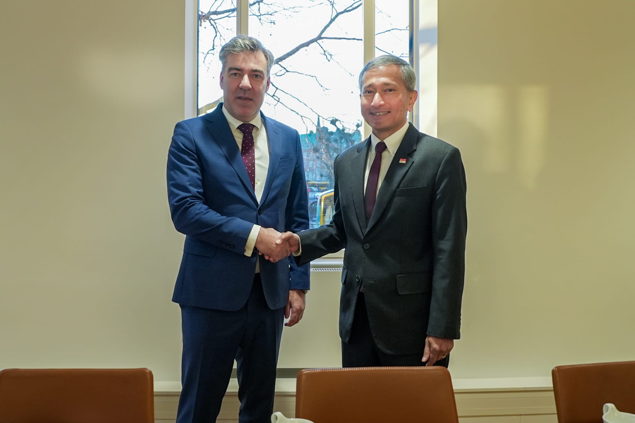 Two men in suits shaking hands in front of a window.